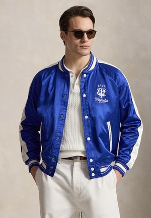 Uomo che indossa occhiali da sole scuri, una giacca di raso blu con strisce bianche e logo "Wimbledon Polo 1877", maglia bianca a maglia e pantaloni bianchi con le mani nelle tasche.