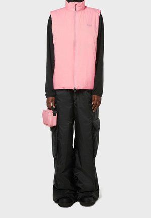 Rosa isolierte Weste mit hohem Kragen, über einem schwarzen Langarmshirt getragen. Schwarze weite Hose mit Taschen. Kleine rosa Tasche in der Hand gehalten.