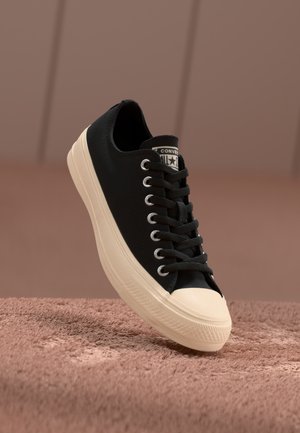 Sorte kanvas sneakers med kremfarget gummitå, snøring, sølvfargede hull og teksturert såle, hviler på en myk, rosa bakgrunn.