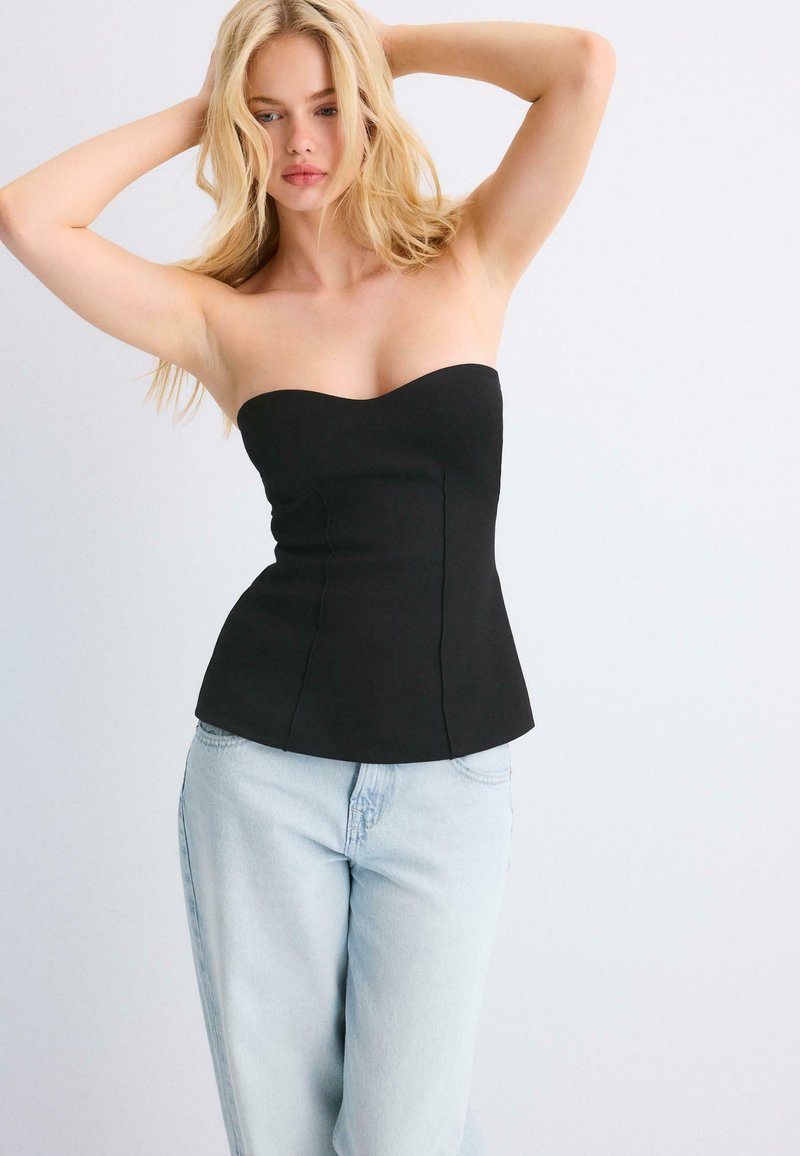 Femme blonde debout, les mains derrière la tête, portant un haut noir sans bretelles et un jean bleu clair, sur un fond uni.