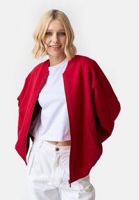 Czerwona kurtka bomberka z zamkiem, prążkowanym kołnierzem i luźnym krojem. Nosiwana na białej krótkiej koszulce i zestawiona z jasnymi spodniami.