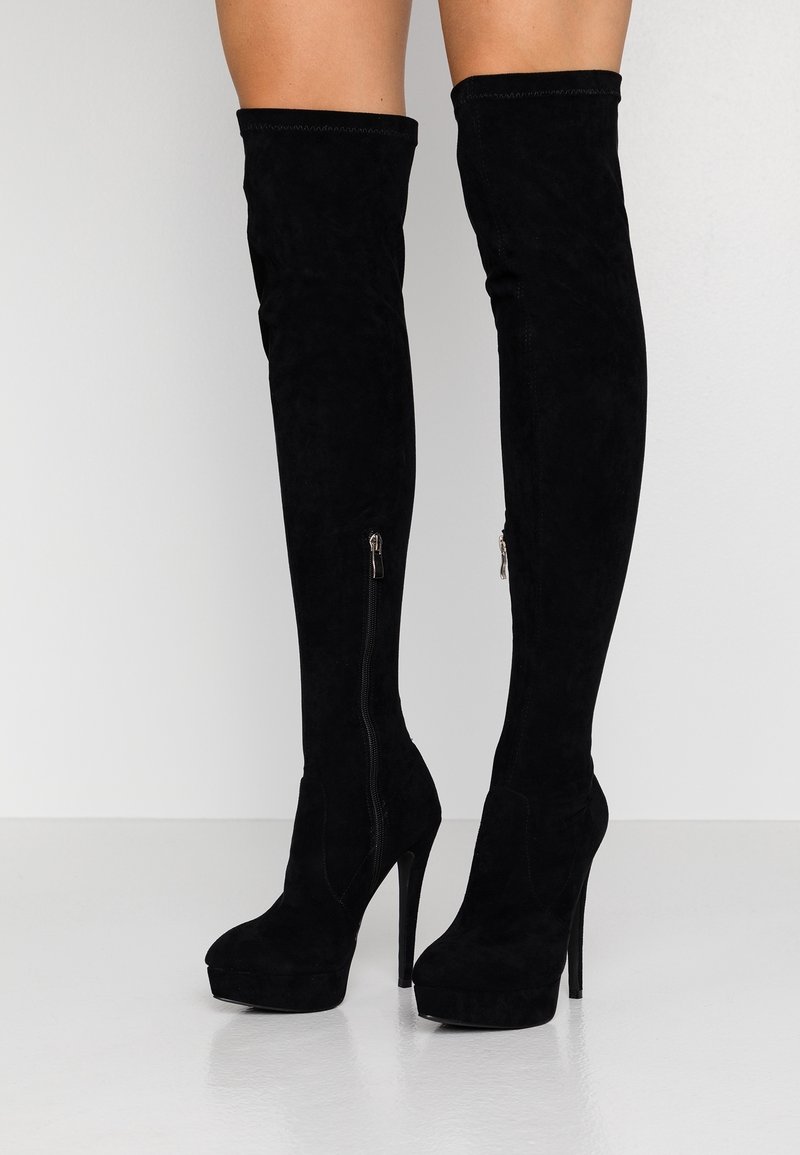 Bottes hautes en daim noir jusqu'à la cuisse avec plateforme, talons aiguilles et fermetures éclair latérales, portées sur des jambes nues sur un fond uni.