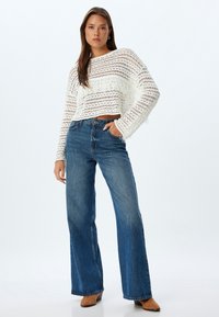 Pull cropped blanc en maille avec des franges, associé à un jean bleu taille haute à jambes larges. Le mannequin porte des chaussures beiges.