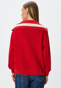 Czerwony sweter z pr�ążkowanym białym i beżowym kołnierzem. Miękka tekstura, luźny fason oraz prążkowane mankiety i dół. Widok z tyłu pokazujący wzór na ramionach.