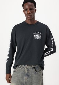 Schwarzes langärmliges Shirt mit weißen Grafiken, darunter "OBEY" auf der Brust und "NEW VISIONS" entlang der Ärmel, kombiniert mit hellen Jeans.