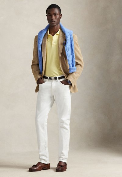 Καφέ σακάκι με ψαροκόκαλο, κίτρινη πόλο μπλούζα, λευκό τζιν, ανοιχτό μπλε πουλόβερ περασμένο στους ώμους, καφέ δερμάτινα λοφάρια, διακριτικό ρολόι χειρός.