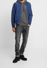 Granatowa kurtka bomberka o gładkim wykończeniu, pod nią szara bluza z kapturem, czarne dżinsy i czarne botki, stojące na neutralnym tle.