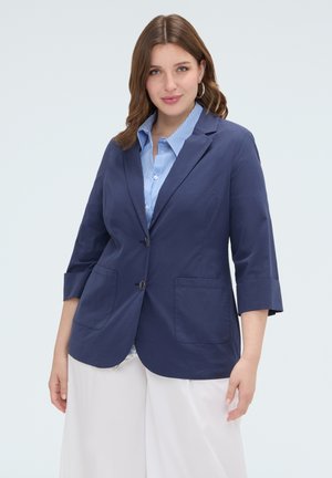 Donna con capelli castani con blazer blu scuro sopra una camicia a righe blu e pantaloni bianchi, in piedi davanti a uno sfondo chiaro uniforme.