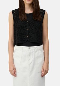 Zwarte mouwloze gehaakte vest met hexagonale patronen, voorzien van knopen aan de voorkant en een gegolfde zoom, gedragen over een zwart topje en een witte rok.