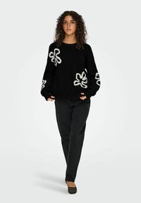 Zwarte gebreide trui met witte geborduurde bloemenmotieven op de mouwen, gecombineerd met zwarte broek en zwarte studded ballerina's, tegen een neutrale achtergrond.