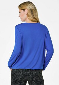 Blaues Langarmoberteil mit rundem Halsausschnitt, weichem Textil und lässiger Passform, kombiniert mit schwarzen gemusterten Hosen.