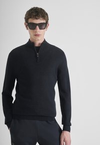 Suéter negro con cremallera en el cuello y mangas largas, hecho de material suave, con detalles texturizados en los hombros y un dobladillo acanalado. Combinado con pantalones oscuros.