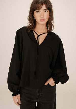 Femme portant un chemisier noir ample à manches longues avec des poignets froncés et un col en V, associé à un jean noir taille haute, se tenant debout avec une main dans la poche.