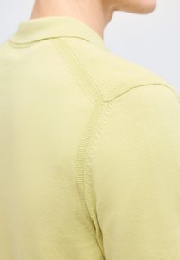 Κοντινό πλάνο ενός ατόμου που φοράει ανοιχτοπράσινο πλεκτό πουλόβερ, με έμφαση στις υφασμάτινες λεπτομέρειες στους ώμους και τον γιακά.