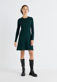 Μακρυμάνικο, πράσινο φόρεμα με εφαρμοστό κορμό και φουσκωτό φούστα; συνδυασμένο με μαύρες μπότες έως το γόνατο; λεία υφή, μίνιμαλ σχέδιο.