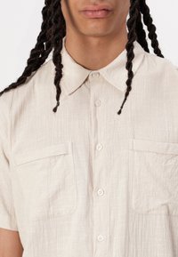 Camisa de manga corta color beige hecha de tela texturizada, con collar, dos bolsillos frontales y cierre de botones con botones blancos.