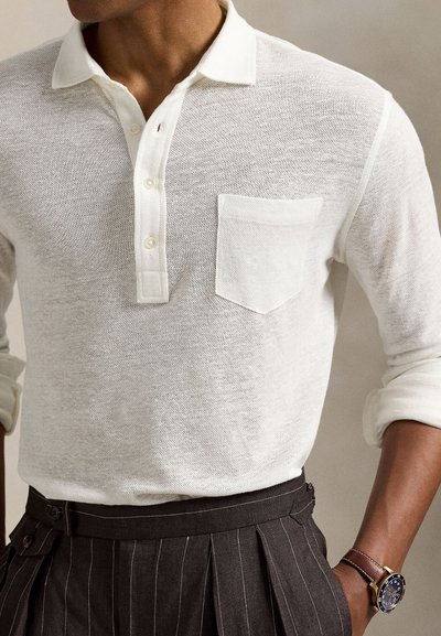Άσπρο πόλο με υφή, με κουμπωτό άνοιγμα, μικρή τσέπη στο στήθος και ρεβέρ στα μανίκια, συνδυασμένο με σκούρο παντελόνι με λεπτές ρίγες και ρολόι.