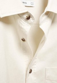 Primo piano di una camicia beige con bottoni, che mostra il colletto, i bottoni superiori e parte della tasca sul petto.
