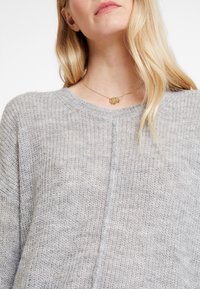 Jasnoszary wełniany sweter z okrągłym dekoltem i luźnym krojem; tekstura jest miękka i ma lekko przezroczystą jakość. Złoty okrągły naszyjnik z zawieszką.