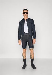 Men's athletic outfit featuring a black lightweight jacket, white mesh top, black cycling shorts, and black cycling shoes with tattoos visible on legs.