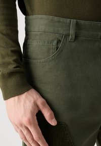 Olive green pants with a textured fabric, featuring a side pocket, belt loops, and a ribbed detail on the leg.