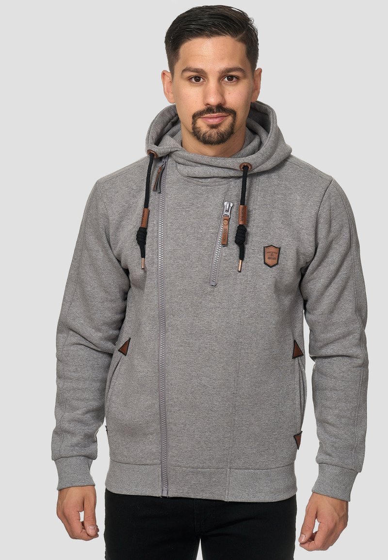 Haut gris à capuche avec fermeture éclair, présentant des accents en cuir marron, des cordons de serrage et des panneaux latéraux triangulaires. Fabriqué en tissu doux et texturé.