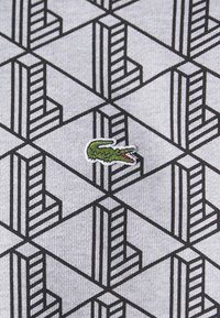 Tessuto grigio con un motivo geometrico nero caratterizzato da linee incrociate. Un piccolo logo di un coccodrillo verde è ricamato sulla superficie.