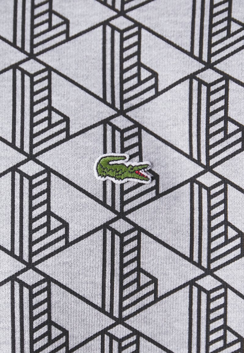 Grauer Stoff mit einem geometrischen schwarzen Muster, das sich kreuzende Linien zeigt. Ein kleines grünes Krokodil-Logo ist auf die Oberfläche gestickt.