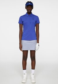 Blå polo med kort ärm, vit mönstrad kjol, vita sneakers och en matchande blå visor. Har en golfhandske på vänster hand.