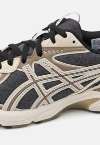 Black and gray athletic sneaker featuring a textured fabric upper, beige accents, black laces, and a cushioned sole with detailed side patterns.