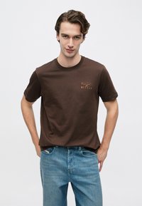 Tricou maro din bumbac, cu mâneci scurte și gât rotund, având un imprimeu subtil cu logo portocaliu pe piept. Blugi albaștri din denim cu croială clasică.