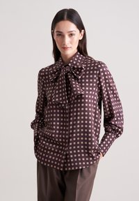Langärmlige, braune Bluse mit einer großen Schleife am Kragen, die mit einem Karomuster aus kleinen cremefarbenen Quadraten und einer glatten Stofftextur gestaltet ist.