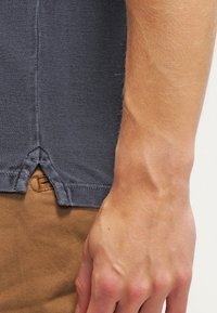 Polo bleu foncé en tissu texturé, avec une fente latérale et un ourlet cousu. Une main repose sur un pantalon marron clair, laissant apparaître le ton de la peau.