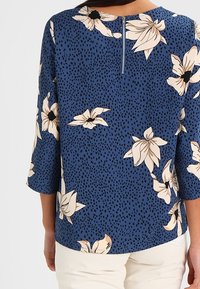 Blouse à fleurs bleues avec manches trois-quarts, dotée d'une fermeture éclair au dos et d'un motif de grandes fleurs crème sur un fond à pois.