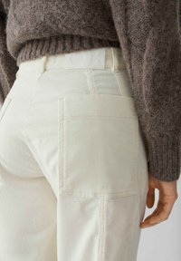 Pantaloni a coste color crema con tasche posteriori, dotati di una fascia in vita bianca con logo, abbinati a un maglione marrone texturizzato.