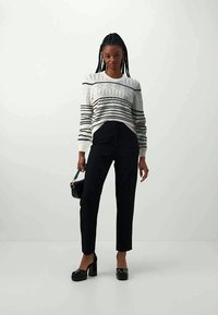 Suéter de malha branco com riscas pretas, combinado com calças pretas ajustadas. A modelo segura uma pequena mala preta e usa sapatos plataforma pretos.