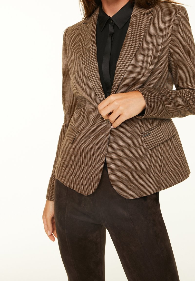 Femme qui boutonne un blazer marron à carreaux porté sur une chemise noire et un pantalon marron foncé dans un décor uni.