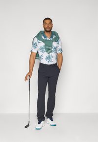 Kortermet blomstret skjorte i lys blå og hvit, marineblå bukser, hvite golf sko, og en grønn genser drapert over skuldrene. Golfklubb i hånden.