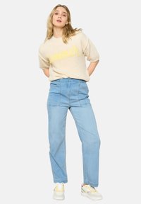 Maglione a maglia beige chiaro con la scritta "Estate" in corsivo giallo. Pantaloni in denim azzurro chiaro a vita alta con taglio dritto. Sneakers bianche con accenti colorati.