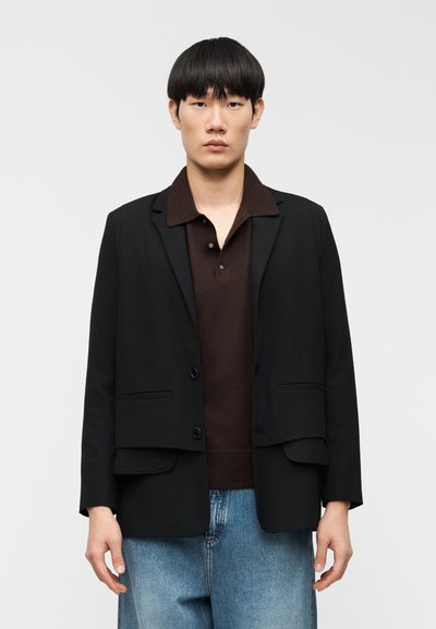 Μαύρο σακάκι με δύο μπροστινές τσέπες, κομψή γραμμή, κλείσιμο με ένα κουμπί· φορεμένο πάνω από καφέ πόλο, σε συνδυασμό με ανοιχτό μπλε φαρδιά τζιν.