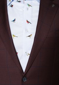 Ciemnoczerwony żakiet z teksturowanym wzorem, nałożony na jasnoniebieską koszulę z kolorowym haftem przedstawiającym ptaki oraz białymi guzikami.