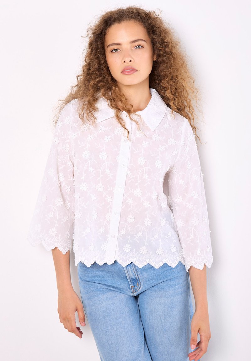 Blusa blanca bordada con dobladillo ondulado, cierre de botones y cuello de pico. Presenta patrones florales y mangas de tres cuartos.