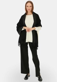 Czarny szal z frędzlami, z ozdobnymi brzegami, noszony na kremowym swetrze, połączony z czarnymi spodniami o szerokich nogawkach i czarnymi butami.