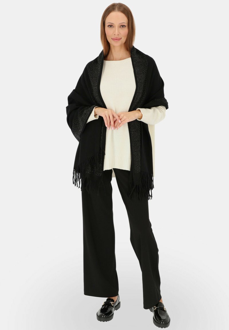 Czarny szal z frędzlami, z ozdobnymi brzegami, noszony na kremowym swetrze, połączony z czarnymi spodniami o szerokich nogawkach i czarnymi butami.