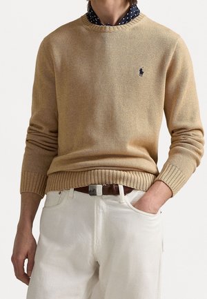 Person trägt beige Strickjacke über einem marineblauen Hemd mit Punkten, weiße Hose und braunen Gürtel, eine Hand in der Tasche, neutraler Hintergrund.