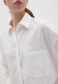 Camisa blanca de lino con botones, que presenta un collar puntiagudo, un bolsillo frontal y una tela texturada. Corte relajado con un diseño minimalista.