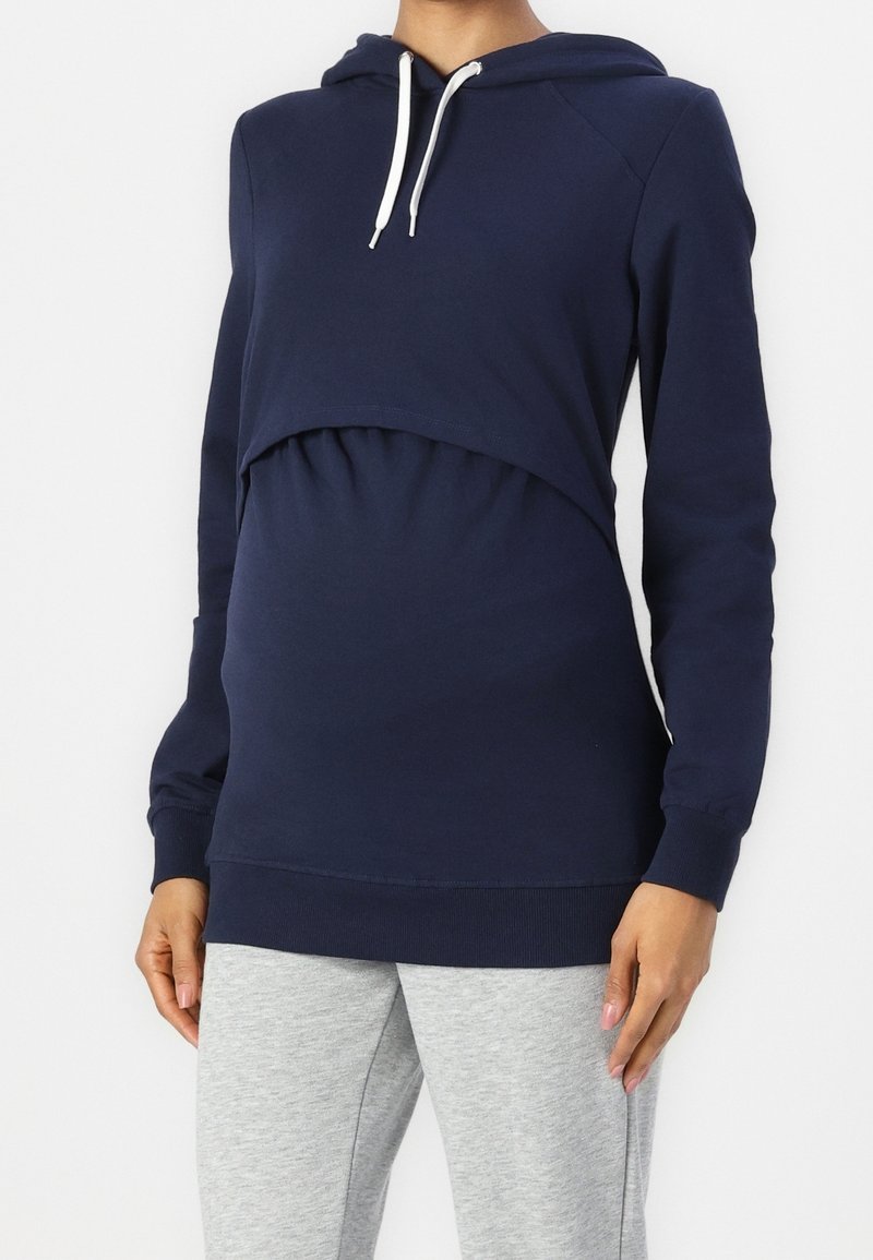 Marineblauwe hoodie met een samengeknepen, gekruiste voorkant, lange mouwen en een capuchon met trekkoord. Gemaakt van zacht materiaal, gecombineerd met grijze sweatpants.