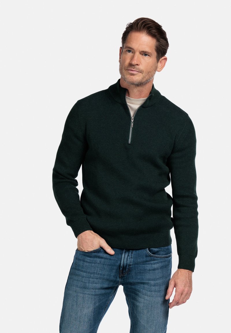 Donkergroen geribde pullovertrui met een halve rits op de kraag. Gedragen over een lichtgekleurde shirt, gecombineerd met blauwe jeans. Geweven stof met textuur.