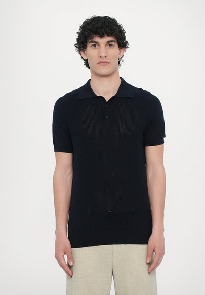 Marineblå kortærmet polo skjorte med et tekstureret strikkemønster, tre-knaps placket og lige kant. Sammen med beige linned bukser.