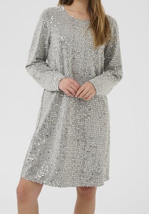 Vrouw draagt een zilveren jurk met lange mouwen, bedekt met reflecterende pailletten, staand met de handen gevouwen over de taille.